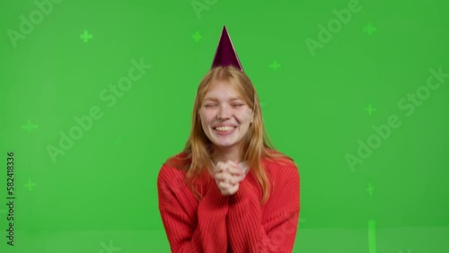 Attractive red-haired white girl in a party hat celebrates her birthday against the backdrop of a green screen. Happy young woman in jeans and knitted jumper blowing out candles on virtual cake photo
