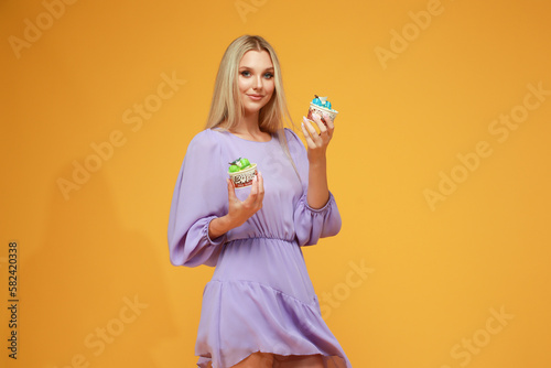 Beautiful blonde girl in a lilac dress with cakes on a yellow background. Studio. Diet and health
