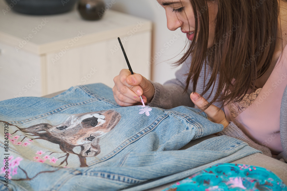 Caucasian woman painting a design of a deer and flowers in a denim jacket.