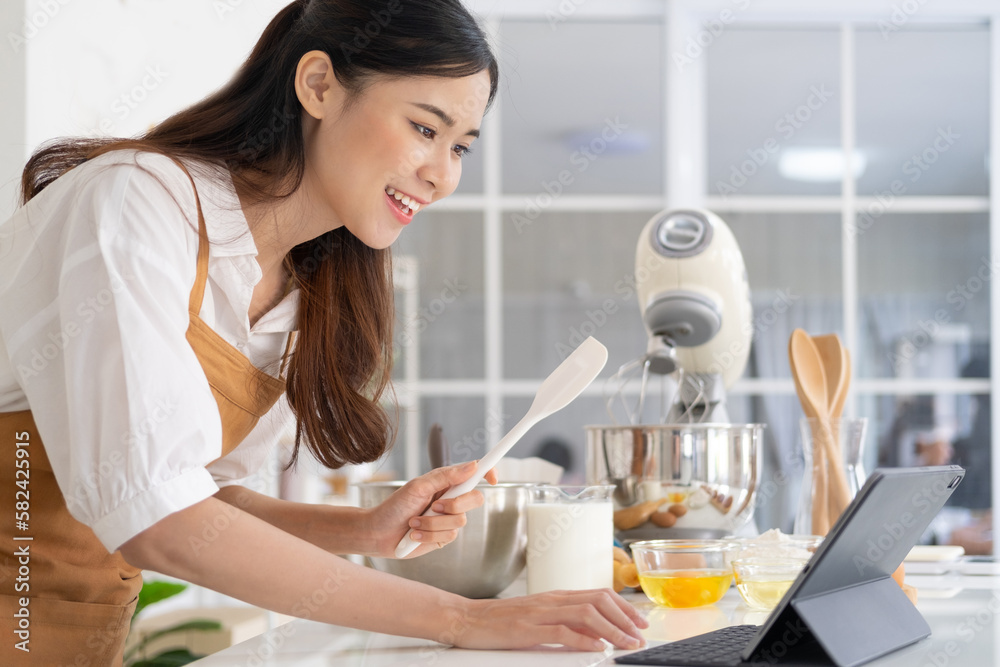 Young asian houes wife learn cooking online. A young woman is watching cooking tutorial video in the kitchen.Reading recipe while making cookies Following Recipe On Digital Tablet. Young asian houes wife learn cooking online. A young woman is watching cooking tutorial video in the kitchen.Reading recipe while making cookies Following Recipe On Digital Tablet.