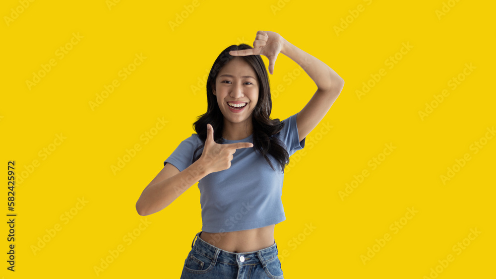 Playful young asian woman in good mood posing for photo or photo frame with fingers on yellow background, Use your fingers to act like a photograph, Mark as if taking a photo. Playful young asian woman in good mood posing for photo or photo frame with fingers on yellow background, Use your fingers to act like a photograph, Mark as if taking a photo.