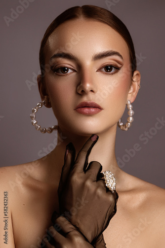 Female model with red hair, brown eyes, freckled skin, poses for a beauty shoot. Model wears pearl hoop earrings. She wears black mesh gloves. Brown background. 