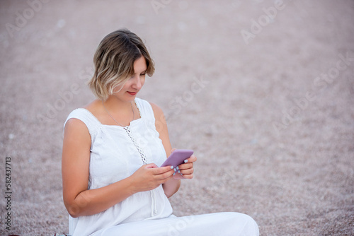 Middle age pregnant woman in white sundress sits in lotus position on the sand by the sea  talking on smartphone. Traveler with backpack  hat. Lifestyle pregnancy tourism concept. Social media