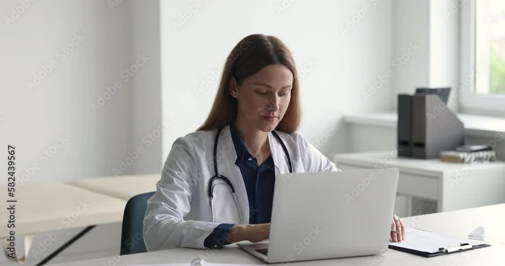 Confident female GP doctor with stethoscope typing on laptop, writing in documents, medical records. Physician woman in white coat working at table in office, using computer Confident female GP doctor with stethoscope typing on laptop, writing in documents, medical records. Physician woman in white coat working at table in office, using computer