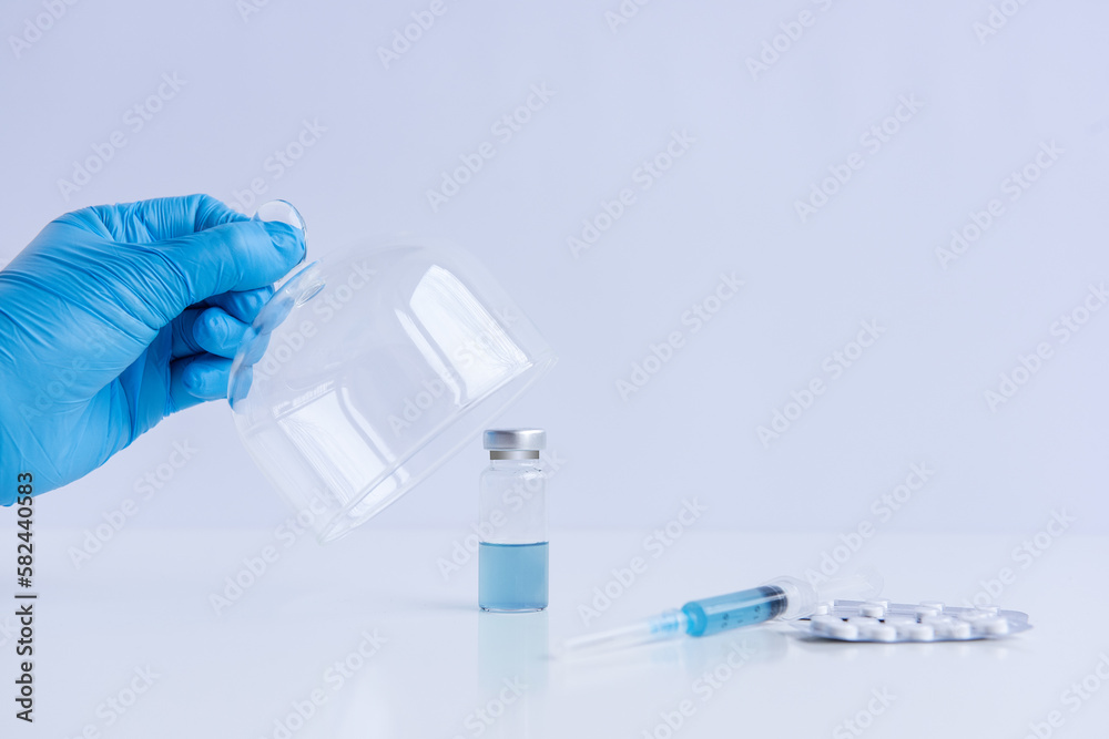 On white isolated background stands the vaccine with syringe, pills. Doctors hand in blue medical glove opens programmed protective dome. Cure against viruses, treatment of people. Mock up, copy space On white isolated background stands the vaccine with syringe, pills. Doctors hand in blue medical glove opens programmed protective dome. Cure against viruses, treatment of people. Mock up, copy space