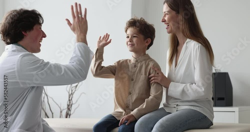 Happy pediatrician man in white coat meeting with cheerful cute little patient boy and mom for examination in office. Male doctor talking to child and parent, laughing, giving high five photo