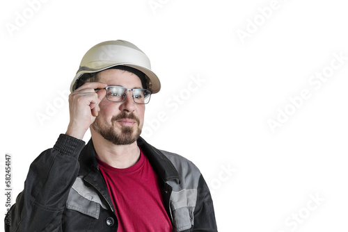 Positive friendly bearded male builder in overalls, hardhat and goggles. Looks at something and maintains glasses. Isolated background.