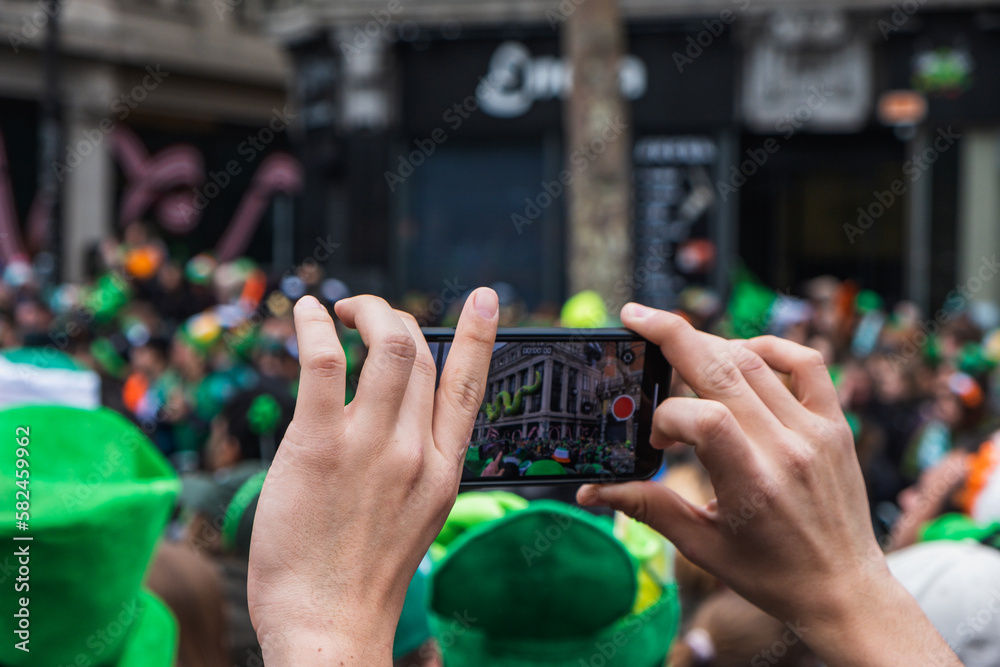 Fototapeta premium Hand with phone, taking picture of the parade, green costume and green hats, people, Paddy's day Dublin centre, Ireland