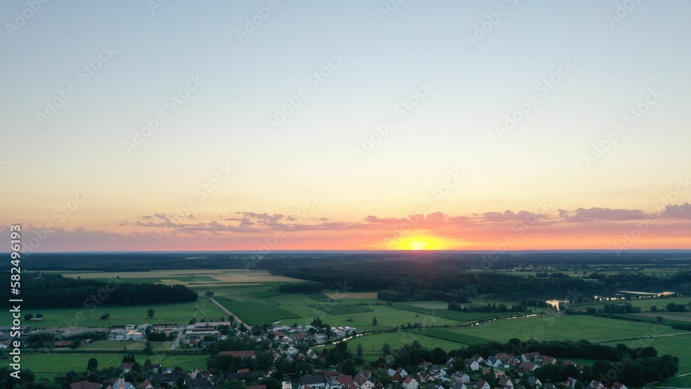 Aerial shot of a small town surrounded by green fields and forests at a beautiful sunset