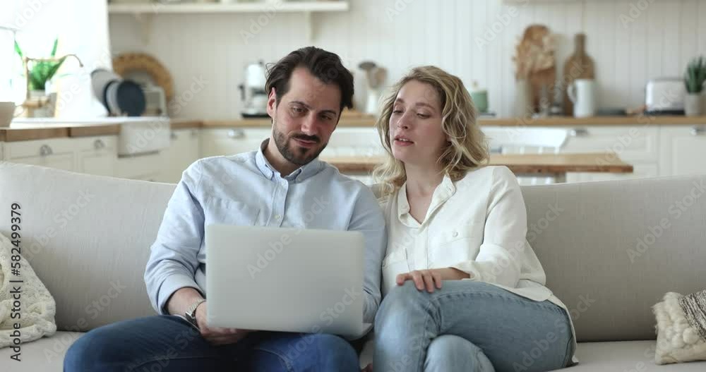 Handsome millennial husband explaining online app work on laptop to wife, showing pc screen, speaking, sitting on couch, getting cheerful, happy, using gadget for home internet communication Handsome millennial husband explaining online app work on laptop to wife, showing pc screen, speaking, sitting on couch, getting cheerful, happy, using gadget for home internet communication