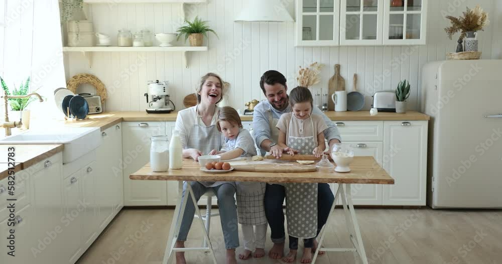 Cheerful loving mom and dad teaching little kids in aprons to cook, baking dessert together, rolling dough at floury table with eggs, milk, talking, laughing. Full length shot, modern kitchen interior Cheerful loving mom and dad teaching little kids in aprons to cook, baking dessert together, rolling dough at floury table with eggs, milk, talking, laughing. Full length shot, modern kitchen interior