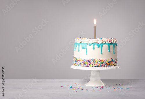 Vanilla Birthday Cake with a lit gold candle, teal blue drip and colorful sprinkles on a light grey white background