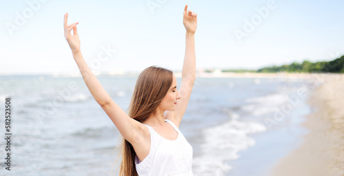 Happy smiling woman in free happiness bliss on ocean beach standing with raising hands. Portrait of a multicultural female model in white summer dress enjoying nature during travel holidays vacation © rogerphoto