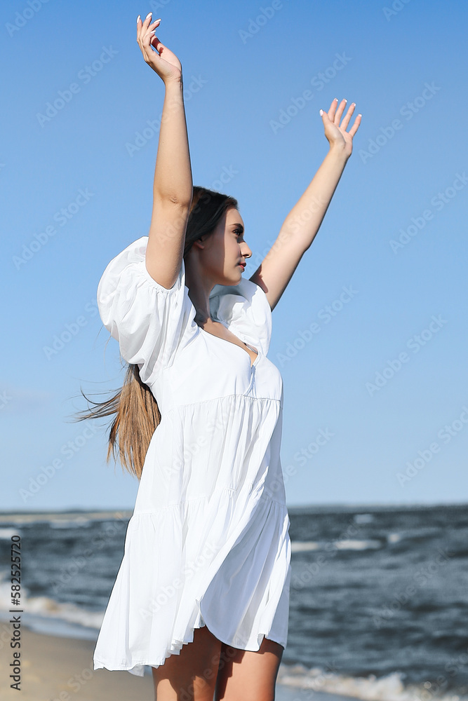 Happy, beautiful woman on the ocean beach standing in a white summer dress, raising hands Happy, beautiful woman on the ocean beach standing in a white summer dress, raising hands