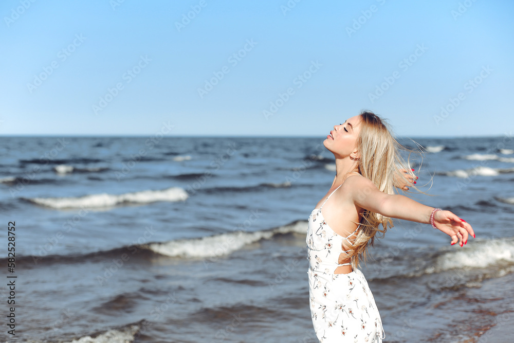 Happy blonde beautiful woman on the ocean beach standing in a white summer dress, open arms