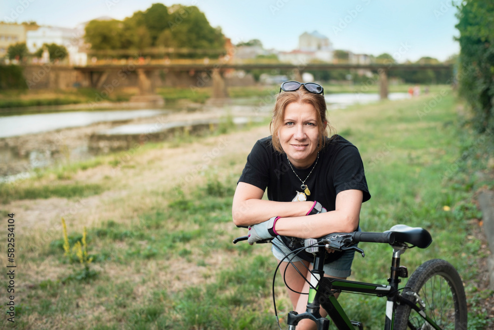 A woman likes active recreation, often rides bicycles together with her husband or friends, now during another ride she rests by the river in the city and smiles A woman likes active recreation, often rides bicycles together with her husband or friends, now during another ride she rests by the river in the city and smiles
