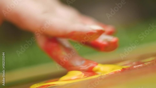 Close-up shot of a finger smearing liquid drops of bright saturated yellow and red acrylic paint on the surface of an abstract background.