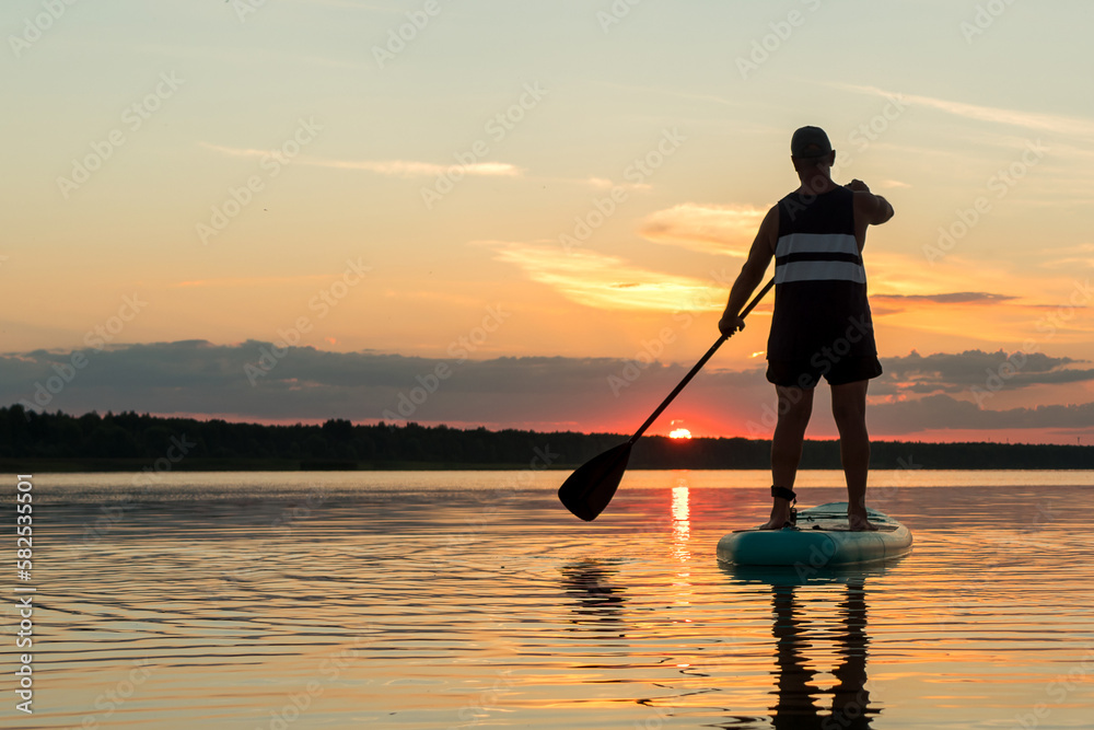 A man in shorts on a sapboard with an oar swims in the lake against the backdrop of the sunset sky.