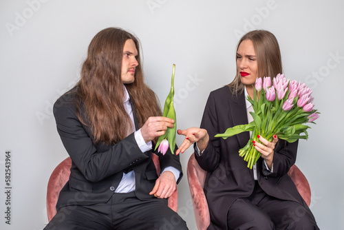 Man with long, brown hair presenting a single tulip to a beautiful lady. Man holding a tulip upside down. Woman expressing disappointmet and holding a bouquet of pink tulips. Serious couple in suits. photo