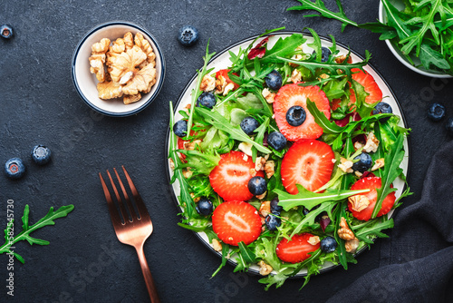 Strawberry and herbs healthy salad with arugula, blueberries and walnuts, black kitchen table, place for text. Fresh useful vegan dish for healthy eating