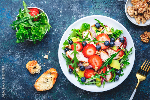 Strawberry, grilled chicken and herbs healthy salad with arugula, blueberries, avocado and walnuts, blue kitchen table. Fresh useful dish for healthy eating