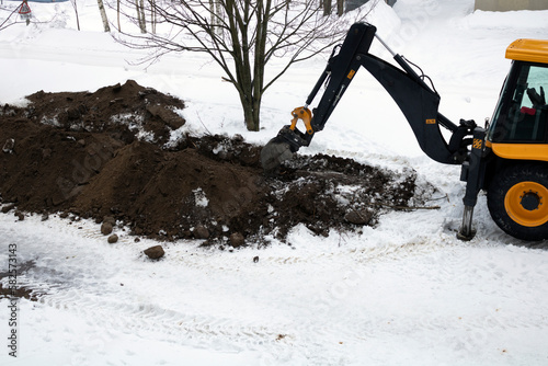 Tractor, excavator digs the earth with a bucket in winter, repairs roads and pipes in the city.