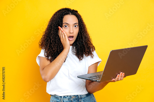 Amazed shocked brazilian or hispanic woman with curly hair, wearing white basic t-shirt, holding an open laptop in hand, looking at the camera in surprised, standing on isolated yellow background