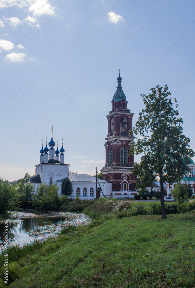 The ancient bell tower of the Intercession Church with a reflection in the river among green trees on a sunny summer day in Yuriev-Polsky russia