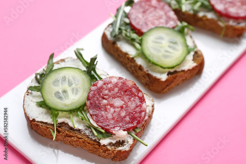 Plate of tasty sandwiches with cream cheese, salami, cucumber and arugula on color background, closeup