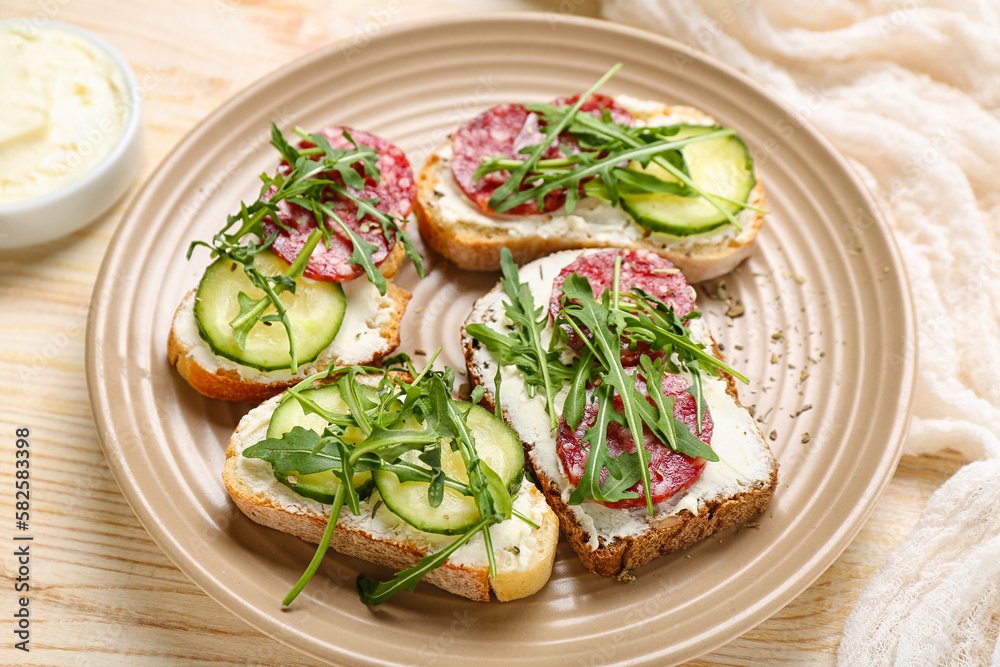 Plate of tasty sandwiches with cream cheese, cucumber, arugula and salami on light wooden table, closeup