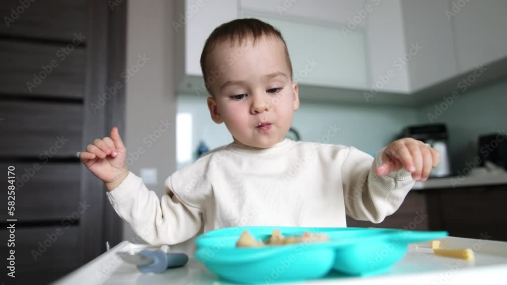 Adorable Caucasian baby in white sweater eating at home. Kid sits at table with blue plate and mom gives a full spoon to her son. Blurred backdrop. Adorable Caucasian baby in white sweater eating at home. Kid sits at table with blue plate and mom gives a full spoon to her son. Blurred backdrop.