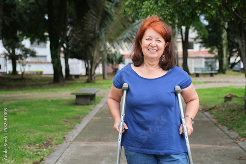 Mature hispanic human recovering from an injury standing with crutches on an urban park pathway, smiling. Concepts: positive attitude towards adversity, physical rehabilitation photo