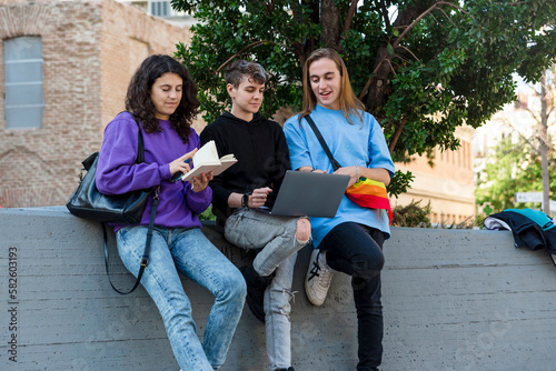 Young diverse people with lgbt rainbow flag using laptop and book outdoors.