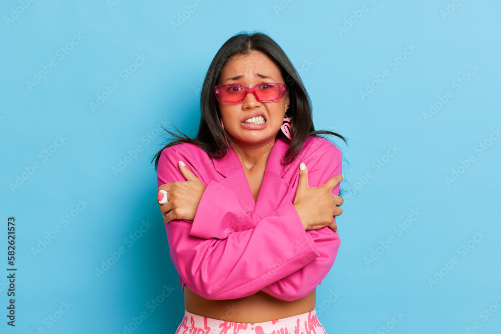 Portrait of young Asian woman in fashionable pink glasses and jacket standing against blue background, she scared of something and hugs herself, copy space, high quality photo Portrait of young Asian woman in fashionable pink glasses and jacket standing against blue background, she scared of something and hugs herself, copy space, high quality photo