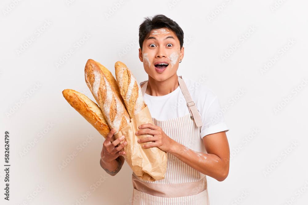 Young Asian man poses on the studio background, smiles holding new-baked baguettes in his hands, happy baker offers his pastries, fresh baking concept, copy space, high quality photo Young Asian man poses on the studio background, smiles holding new-baked baguettes in his hands, happy baker offers his pastries, fresh baking concept, copy space, high quality photo