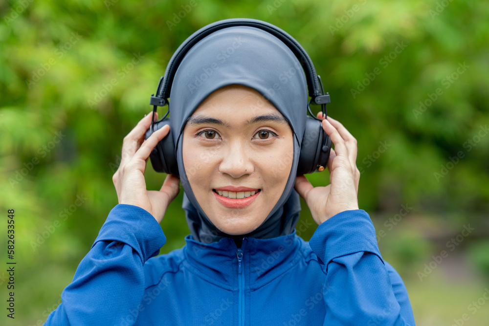 Asian cute muslim woman wearing sportswear and a blue hijab Working out and wearing earphones listening to music on her phone outdoors at the park in the morning with a fresh feeling. Asian cute muslim woman wearing sportswear and a blue hijab Working out and wearing earphones listening to music on her phone outdoors at the park in the morning with a fresh feeling.