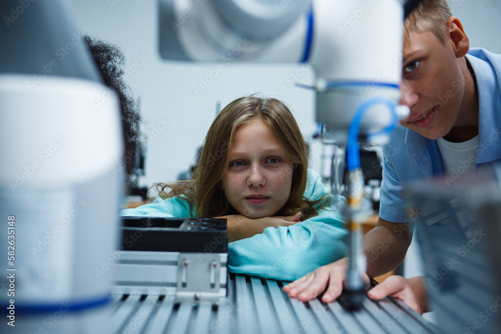 Cute young girl smiling to the camera sitting in front of robot in robotics class in the classroom. Innovation technology and success concept. Cute young girl smiling to the camera sitting in front of robot in robotics class in the classroom. Innovation technology and success concept.