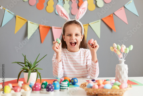 Indoor shot of amazed charming little girl holding small multicolored painted dyed easter eggs, wearing bunny ears looking at camera, screaming with happiness.