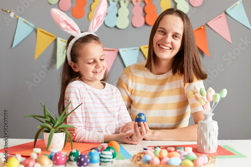 Photo of good looking pretty family preparing for Easter, mother and her daughter painting Easter eggs, little child girl wearing bunny ears, enjoying spending time together.
