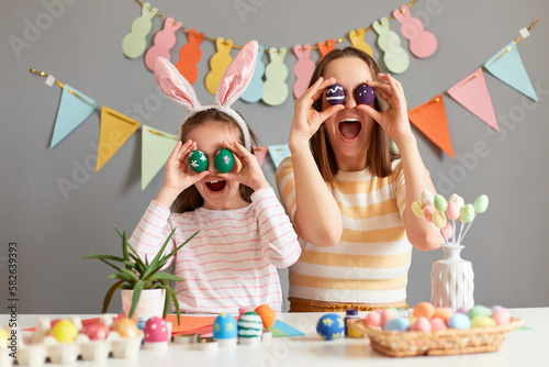 Happy Easter. Image of surprised funny mother and her daughter covering eyes with Easter eggs and screaming with surprised and excitement, little child girl wearing bunny ears © sementsova321