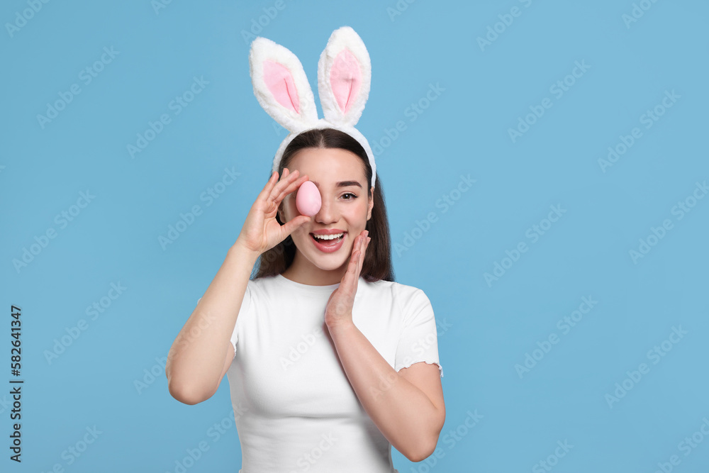 Happy woman in bunny ears headband holding painted Easter egg on turquoise background