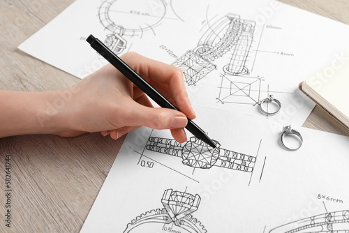 Jeweler drawing sketch of elegant ring on paper at wooden table, closeup