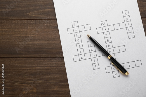 Crossword with answers and pen on wooden table, top view. Space for text