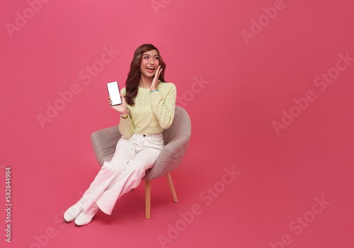 Happy young Asian woman sitting on sofa feeling happiness and gesture showing mobile smartphone blank screen on isolated pink background.