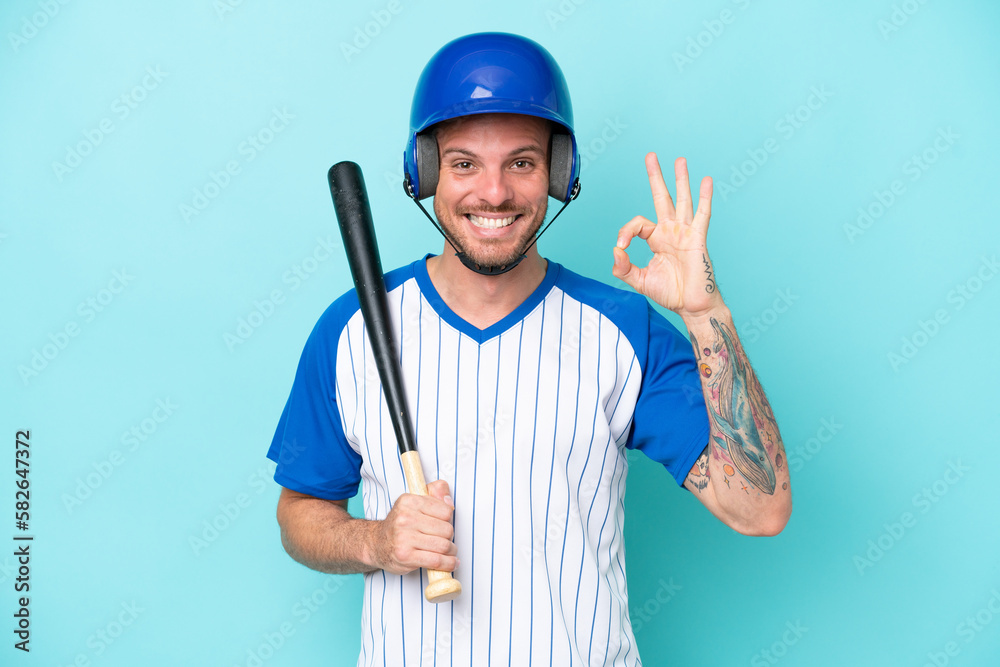 Baseball player with helmet and bat isolated on blue background showing ok sign with fingers