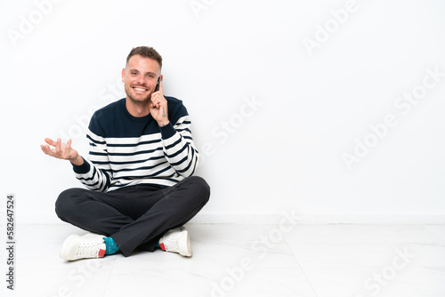 Young man sitting on the floor isolated on white background keeping a conversation with the mobile phone with someone