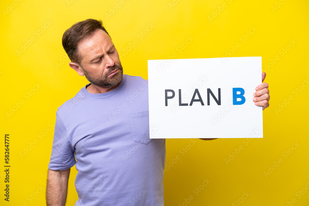 Middle age caucasian man isolated on yellow background holding a placard with the message PLAN B with sad expression Middle age caucasian man isolated on yellow background holding a placard with the message PLAN B with sad expression