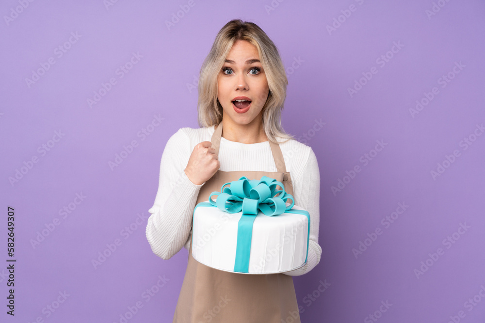 Young pastry chef holding a big cake over isolated purple background celebrating a victory Young pastry chef holding a big cake over isolated purple background celebrating a victory