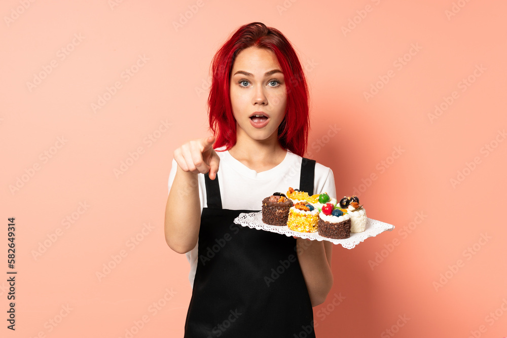 Pastry chef holding a muffins isolated on pink background surprised and pointing front Pastry chef holding a muffins isolated on pink background surprised and pointing front