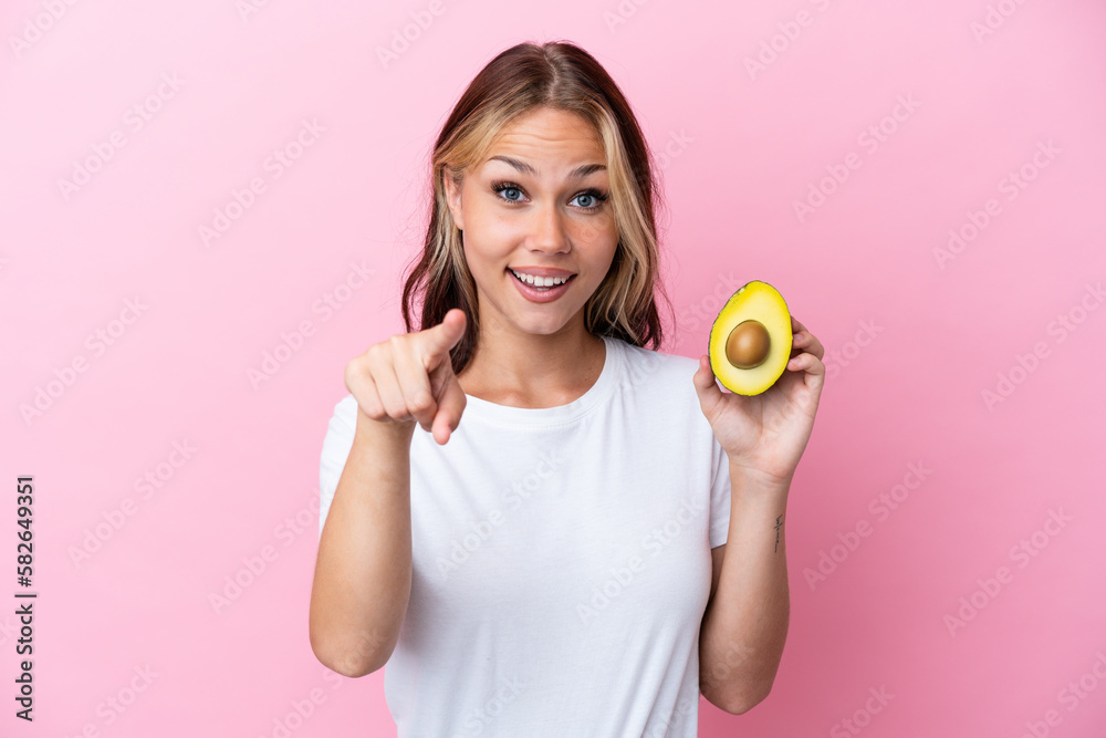 Young Russian woman holding avocado isolated on pink background surprised and pointing front Young Russian woman holding avocado isolated on pink background surprised and pointing front